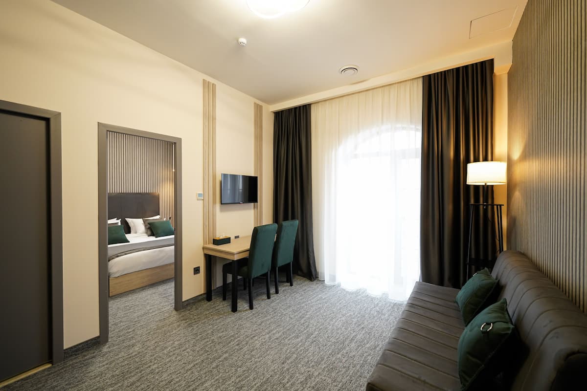 A hotel room with a bed, a desk with chairs, a TV, a sofa, and a lamp.