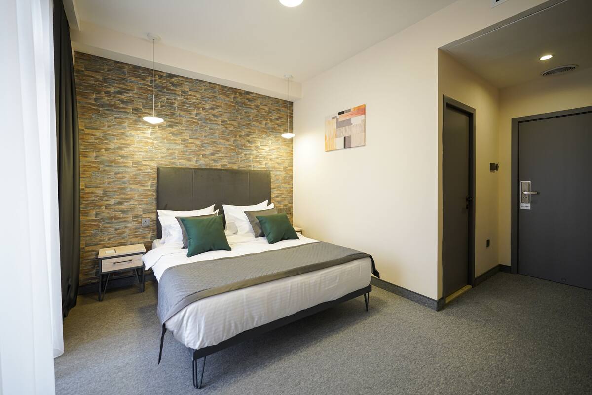 A hotel room with a stone accent wall, a bed with white linens and green pillows, a bedside table, and a door.