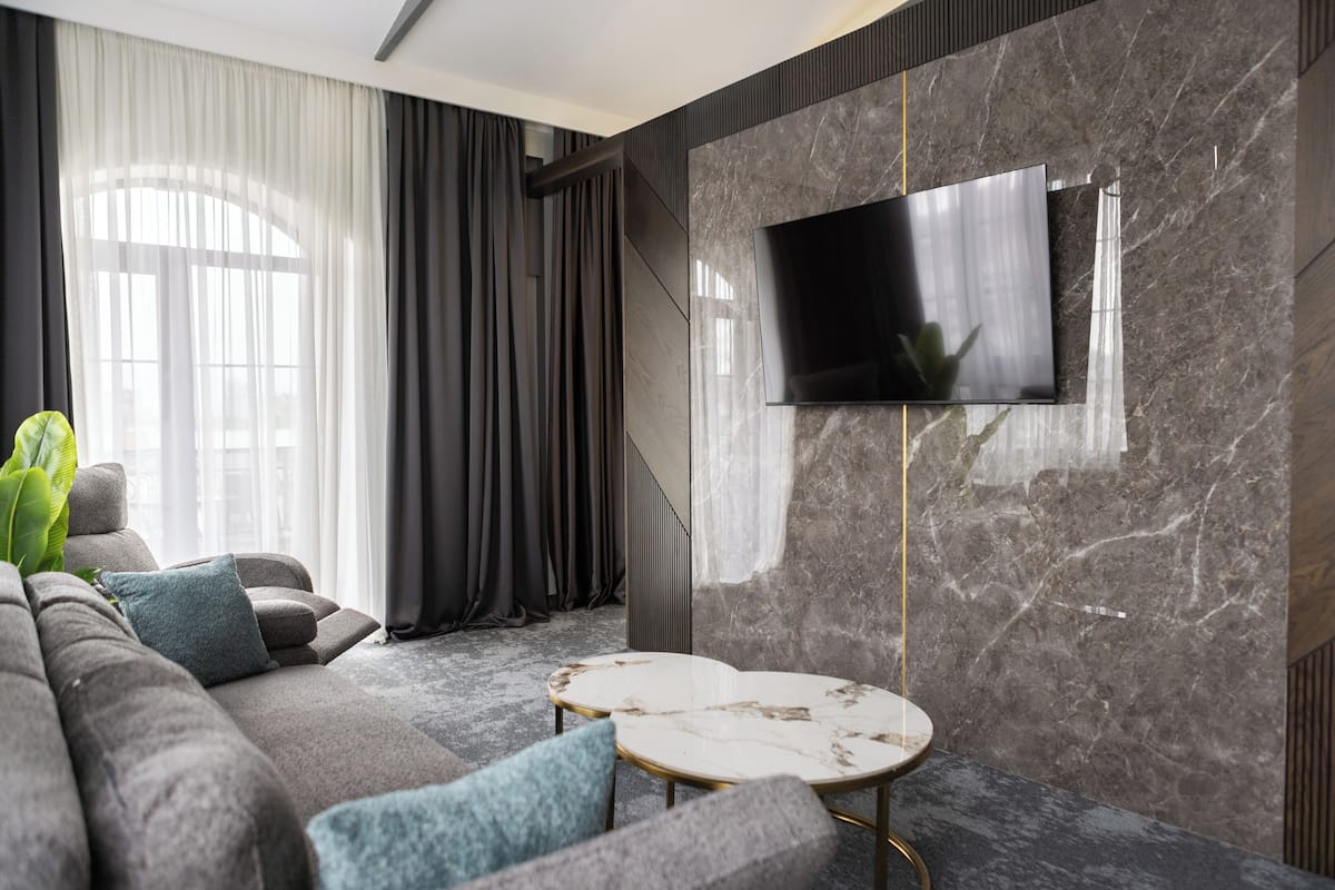 A modern living room with a large flat-screen TV mounted on a marble wall, a grey sofa with blue cushions, and a small round coffee table.