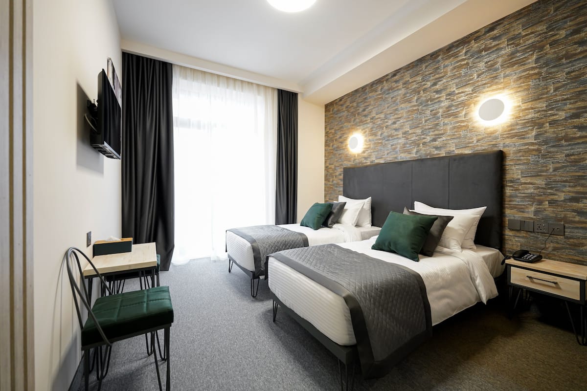 A hotel room with two beds, a desk, a chair, a TV, and a stone wall with lighting.