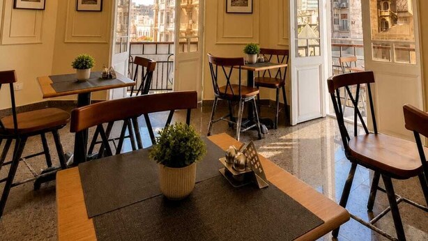 Comedor de hotel con una araña, cuadros enmarcados y vistas de la ciudad a través de amplias ventanas.