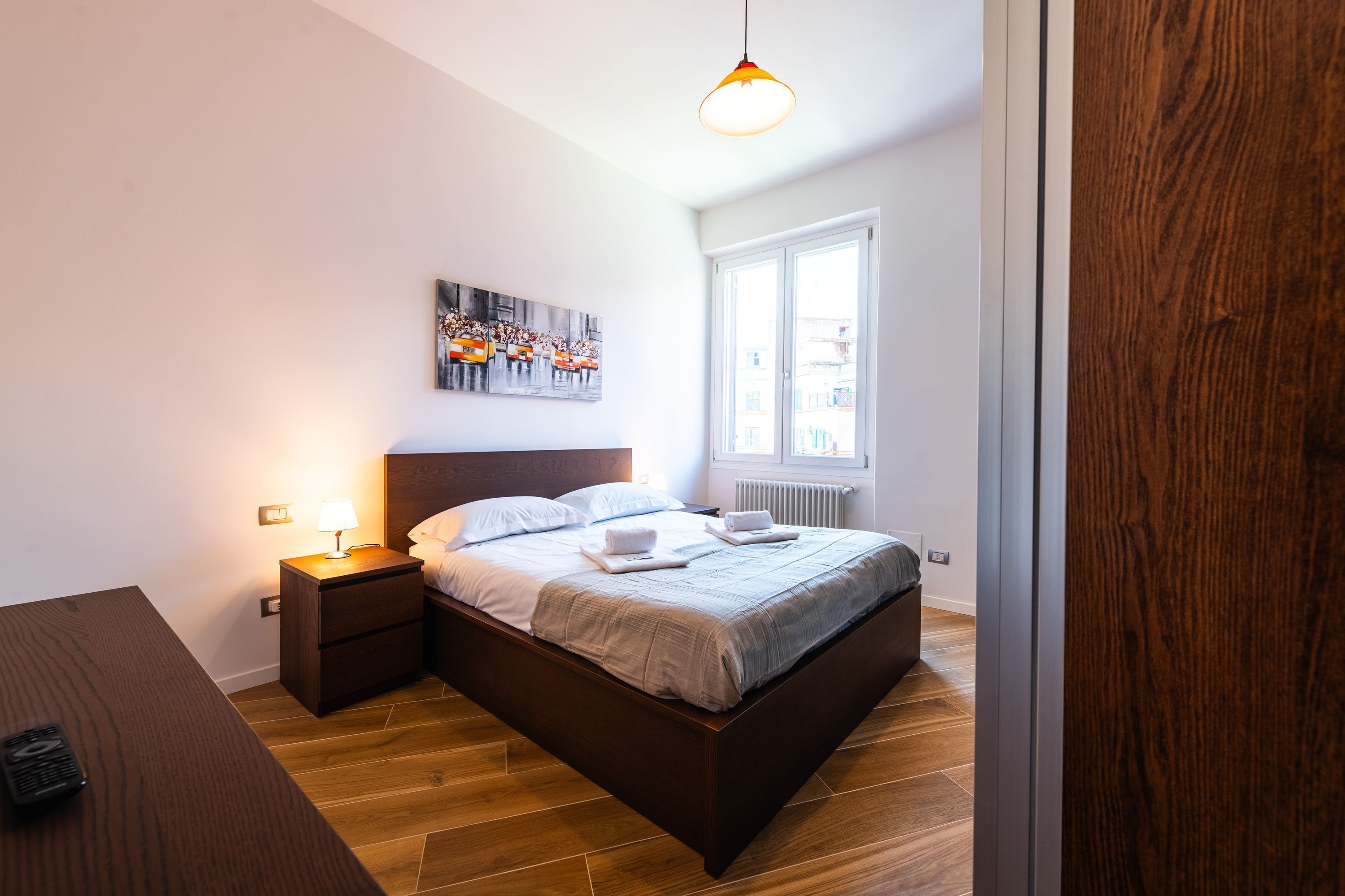Une chambre moderne avec un lit en bois, une table de chevet avec une lampe, un tableau au mur et une fenêtre donnant sur des immeubles.
