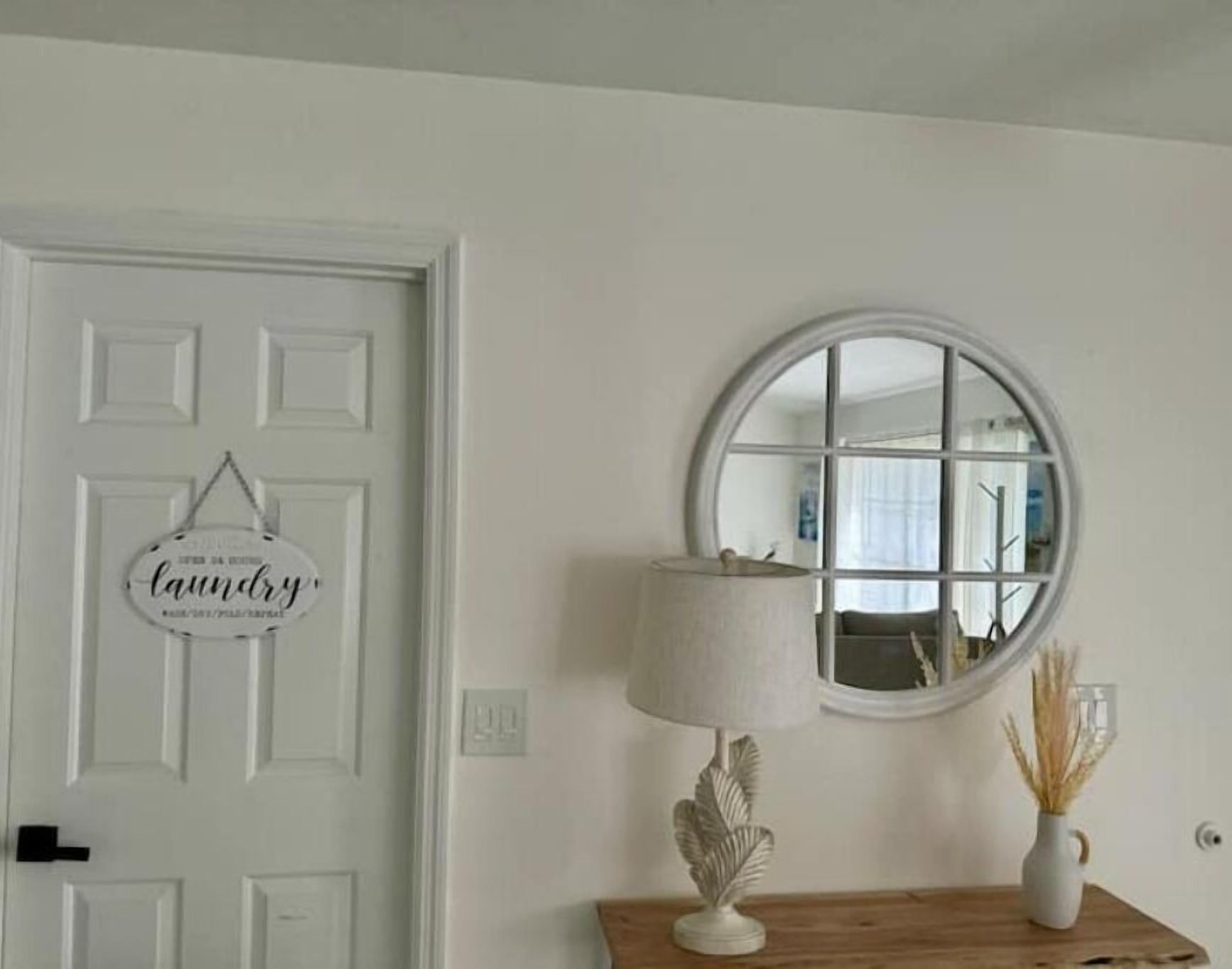 A white door with a 'laundry' sign, a round mirror, a lamp, and a vase with dried plants on a wooden table.