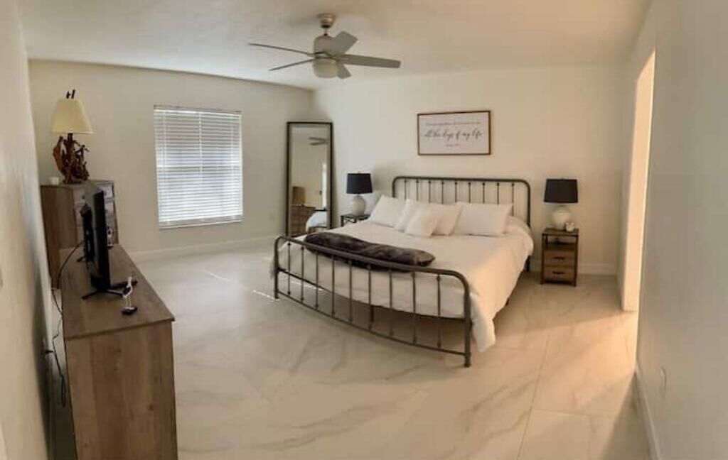 Une chambre à coucher comprenant un lit, des tables de chevet, un ventilateur de plafond, un miroir et un panneau fixé au mur.