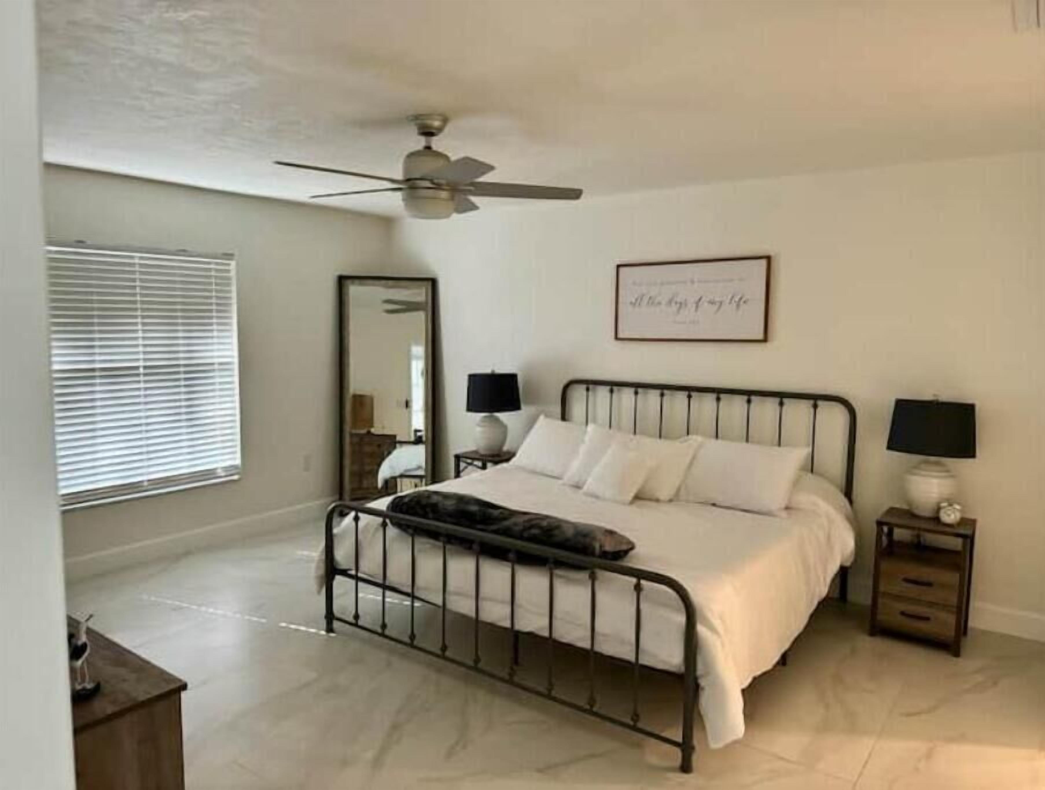 A bedroom with a bed, two bedside tables with lamps, a ceiling fan, a large mirror, and a framed quote on the wall.