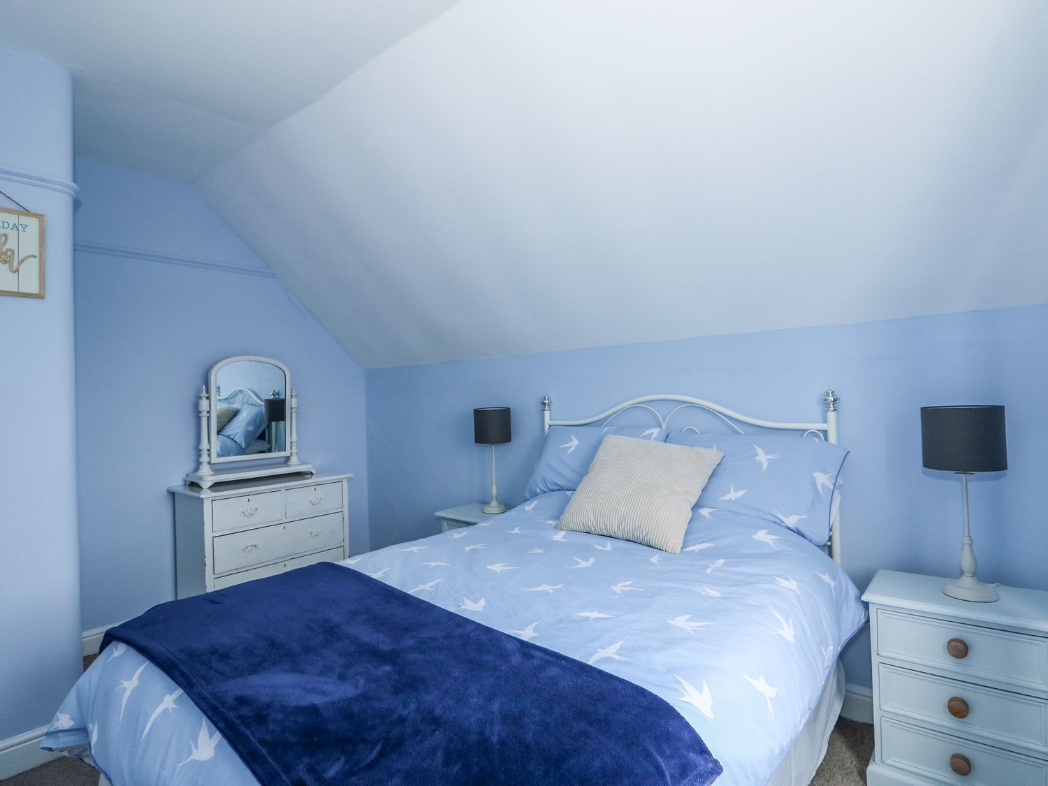 Un dormitorio con techo inclinado, una cama con colcha de diseño azul y blanco, dos mesitas de noche con lámparas, un aparador y un espejo.