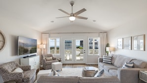 A spacious living room with a ceiling fan, a television, a patterned sofa, a grey sofa, and a view through glass doors to an outdoor area. - Dot's Spot, Almost Oceanfront House in Emerald Isle, NC! (Atlantic Beach)