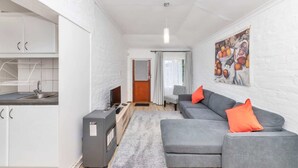 A compact living space with a kitchenette, grey sofa with orange cushions, and a fruit painting on the wall. - Peaceful 1BD Flat w/ Private Gardenrondebosch (Cape Town)