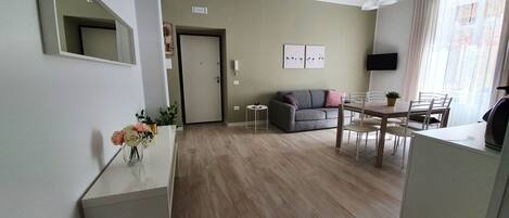 A modern living room with a grey sofa, a dining table with chairs, a small side table, and a wall-mounted TV.
