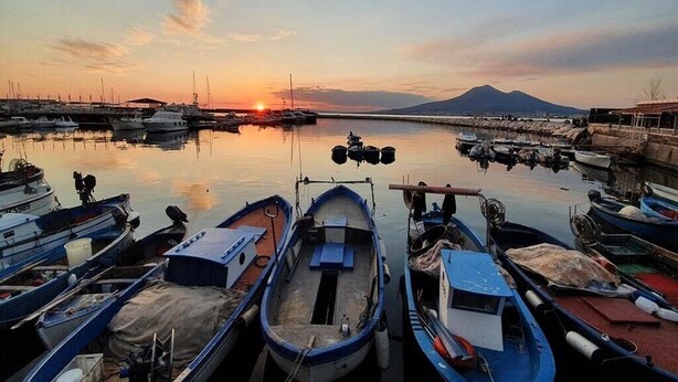 Ein Yachthafen mit anlegenden Booten bei Sonnenuntergang, ein Berg im Hintergrund und ruhiges Wasser, das den Himmel spiegelt.