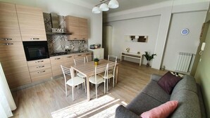 Eine moderne Küche mit Holzschränken, einem Esstisch mit Stühlen und einem grauen Sofa mit einem rosafarbenen Kissen.