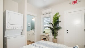 A bedroom with a bed, a washing machine, a mirror, a plant, and an exit sign.