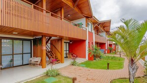 A modern building with a wooden exterior, a covered outdoor seating area, and a landscaped garden with palm trees. - IT57 Apto Pé na Areia - Praia da Espera (Camaçari)