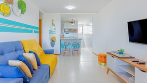 A modern living room with a blue sofa, yellow throw, and a kitchen area in the background. - IT57 Apto Pé na Areia - Praia da Espera (Camaçari)