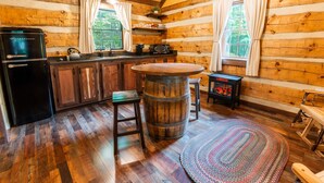 A rustic cabin interior with wooden walls, a wooden bar stool, a round wooden table, a wood-burning stove, and a refrigerator.
