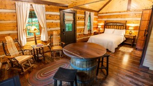A log cabin room with a wooden dining table, two chairs, a bed, a small table with a lamp, and a rug.
