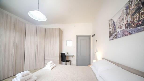 Un dormitorio con cama, escritorio y silla. Se ve una puerta y un cuadro en la pared.