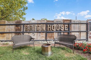 A rustic outdoor seating area with two wicker chairs and a metal fire pit, set against a wooden fence with the words "BRICK HOUSE" visible.