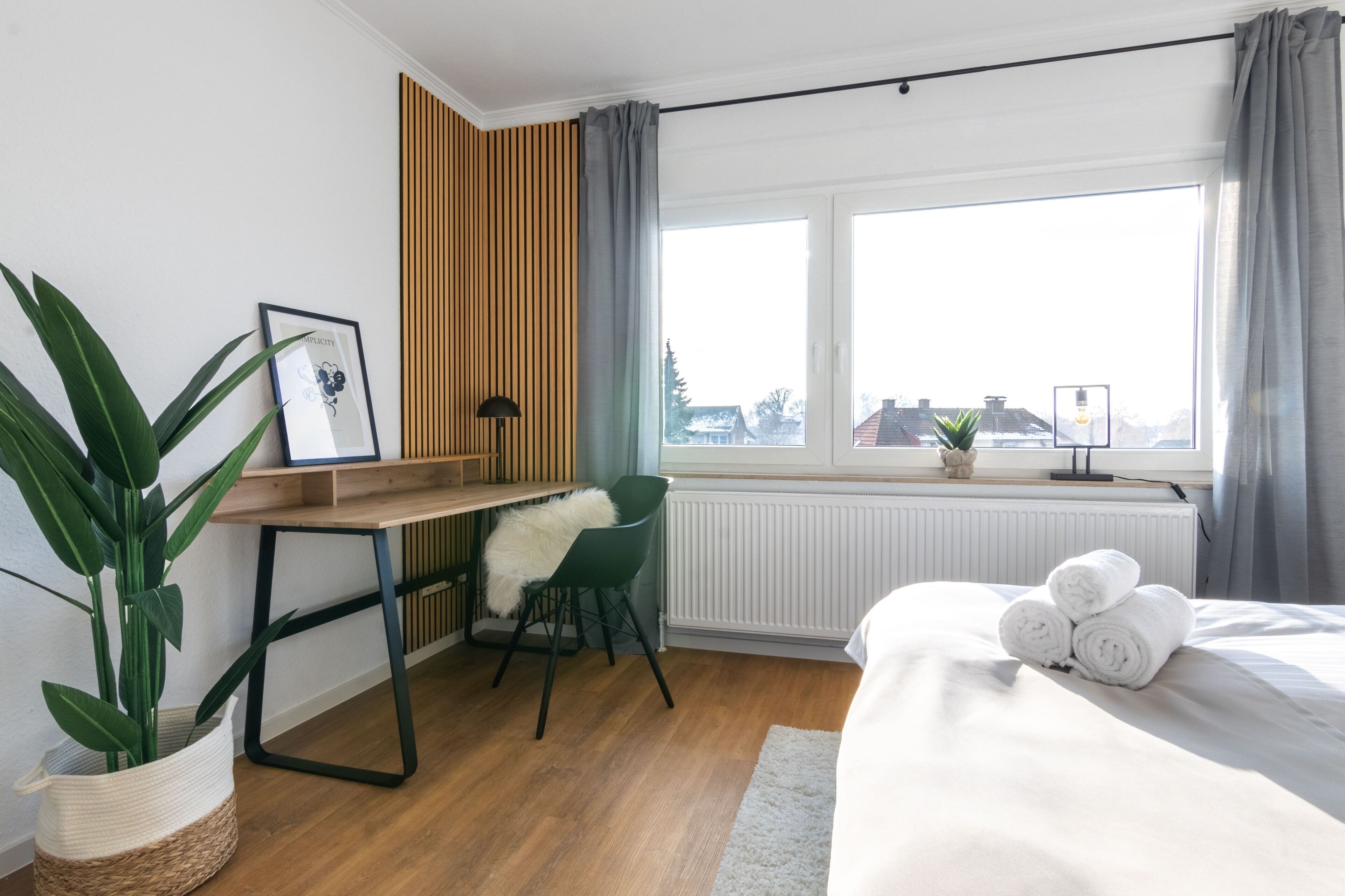 A modern hotel room with a wooden desk, a green chair, a large window with curtains, a bed with rolled towels, and a potted plant.