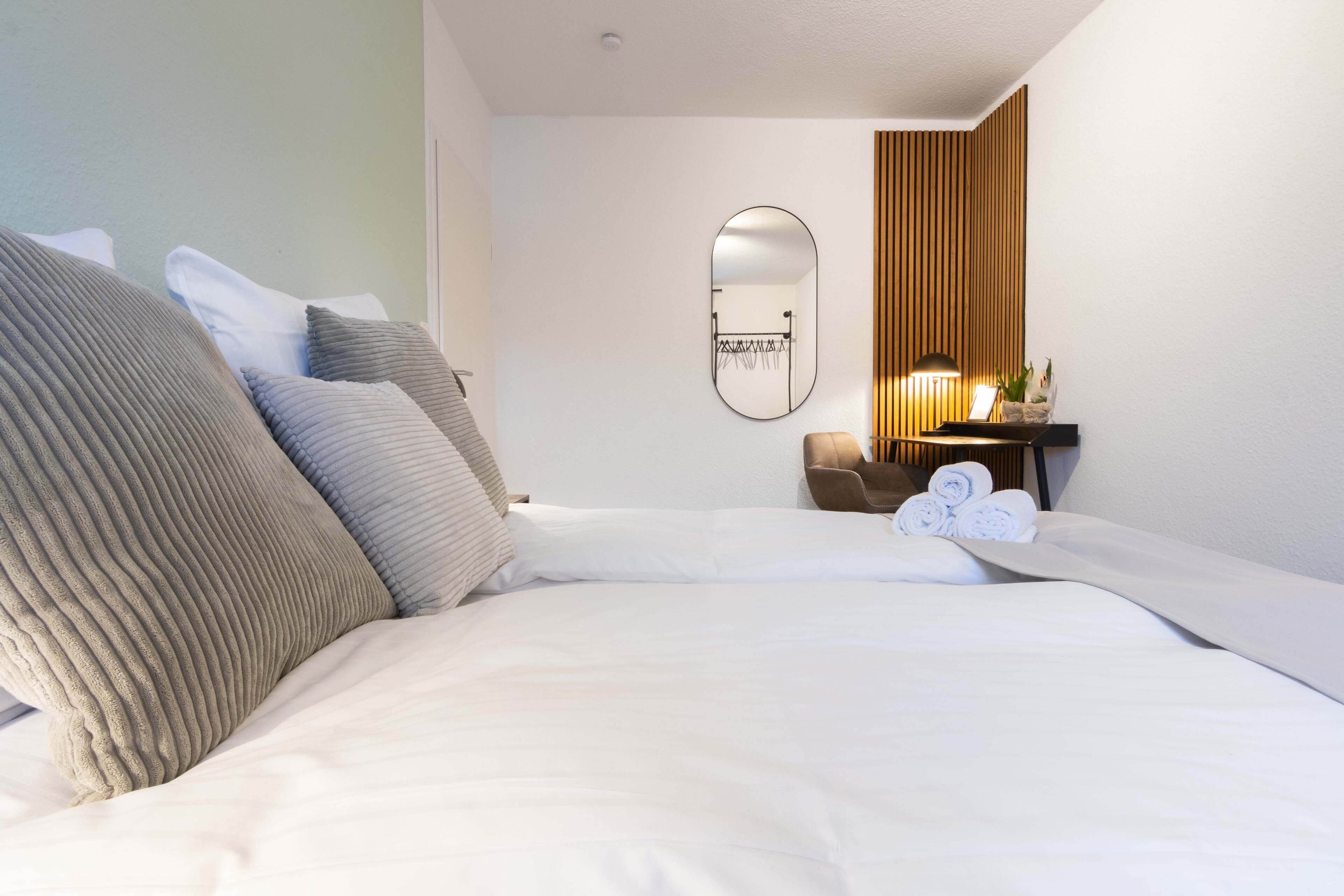 A neatly made bed with white linens and several pillows, a small bedside table with a lamp, and a mirror on the wall.