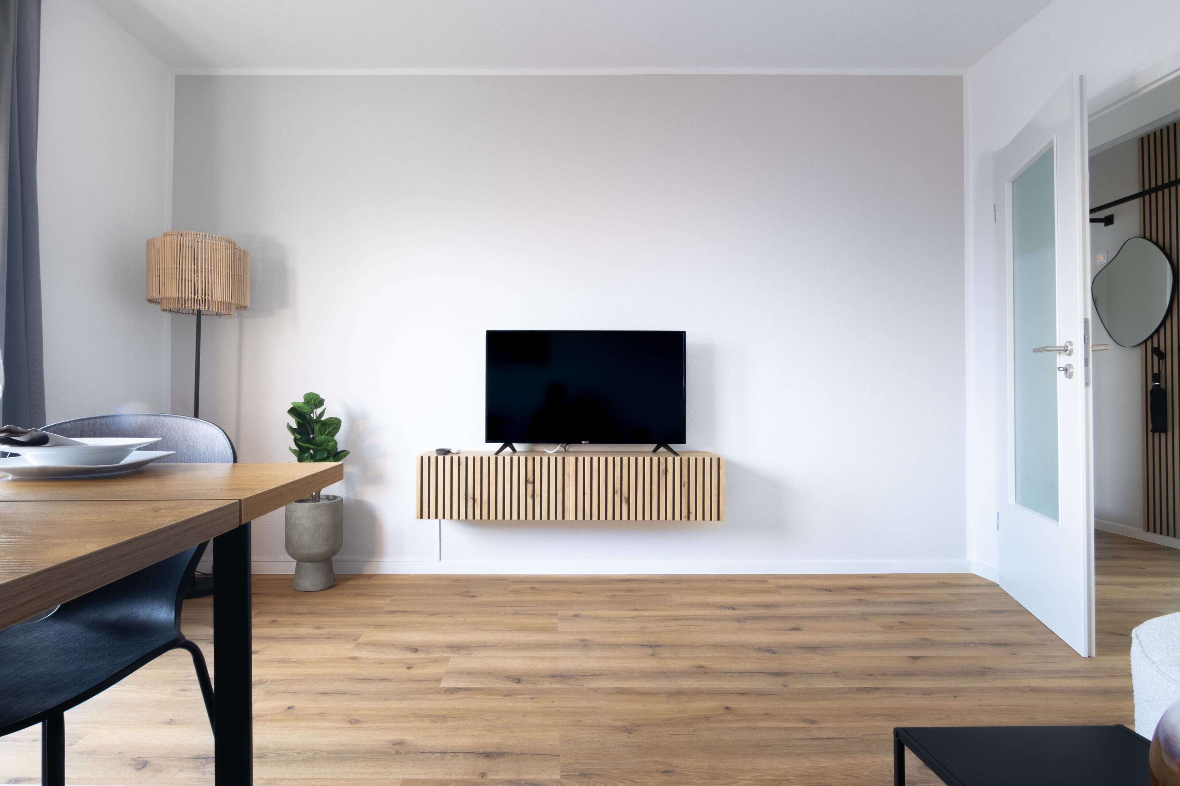Un salón moderno con suelo de madera, un televisor de pantalla plana montado en un panel de madera, una mesa de comedor con sillas, una lámpara de pie y una planta en maceta.