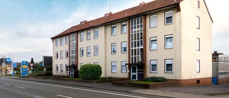 Un edificio de varios pisos con fachada color beige y tejado de tejas rojas. Cuenta con una escalera central, varias ventanas y una pequeña zona de entrada con una puerta.