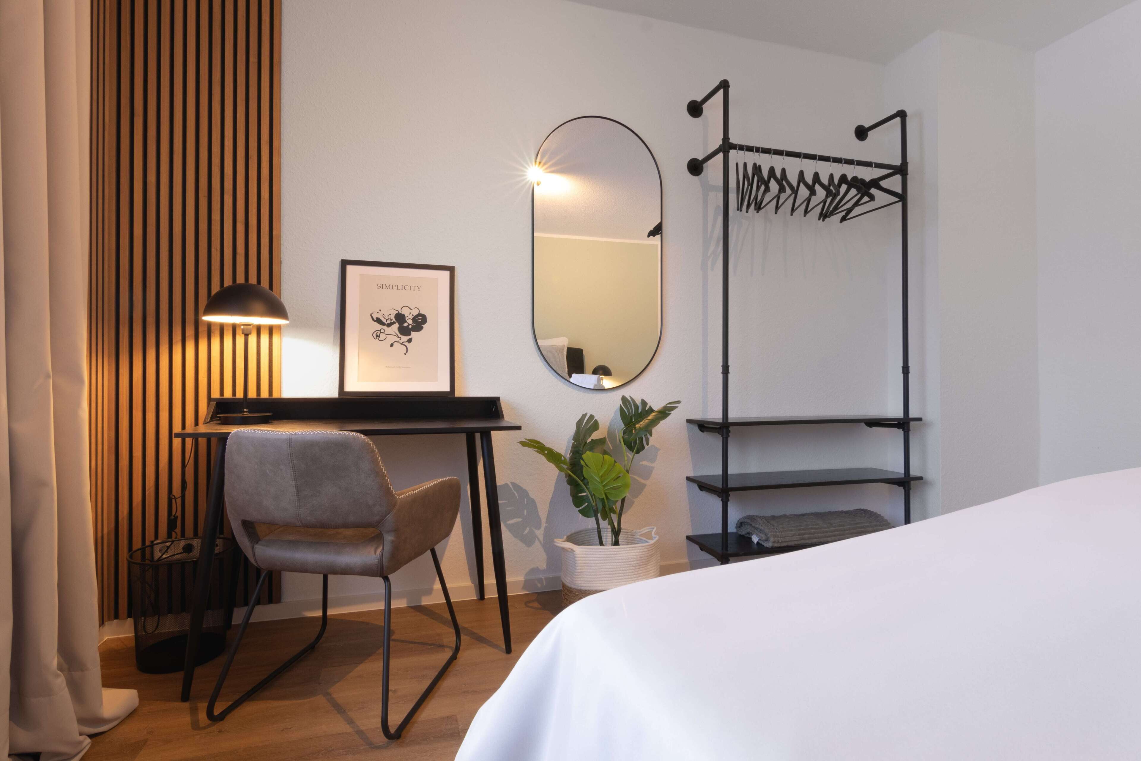 A hotel room with a bed, a desk with a chair, a mirror, a plant, and a drying rack.