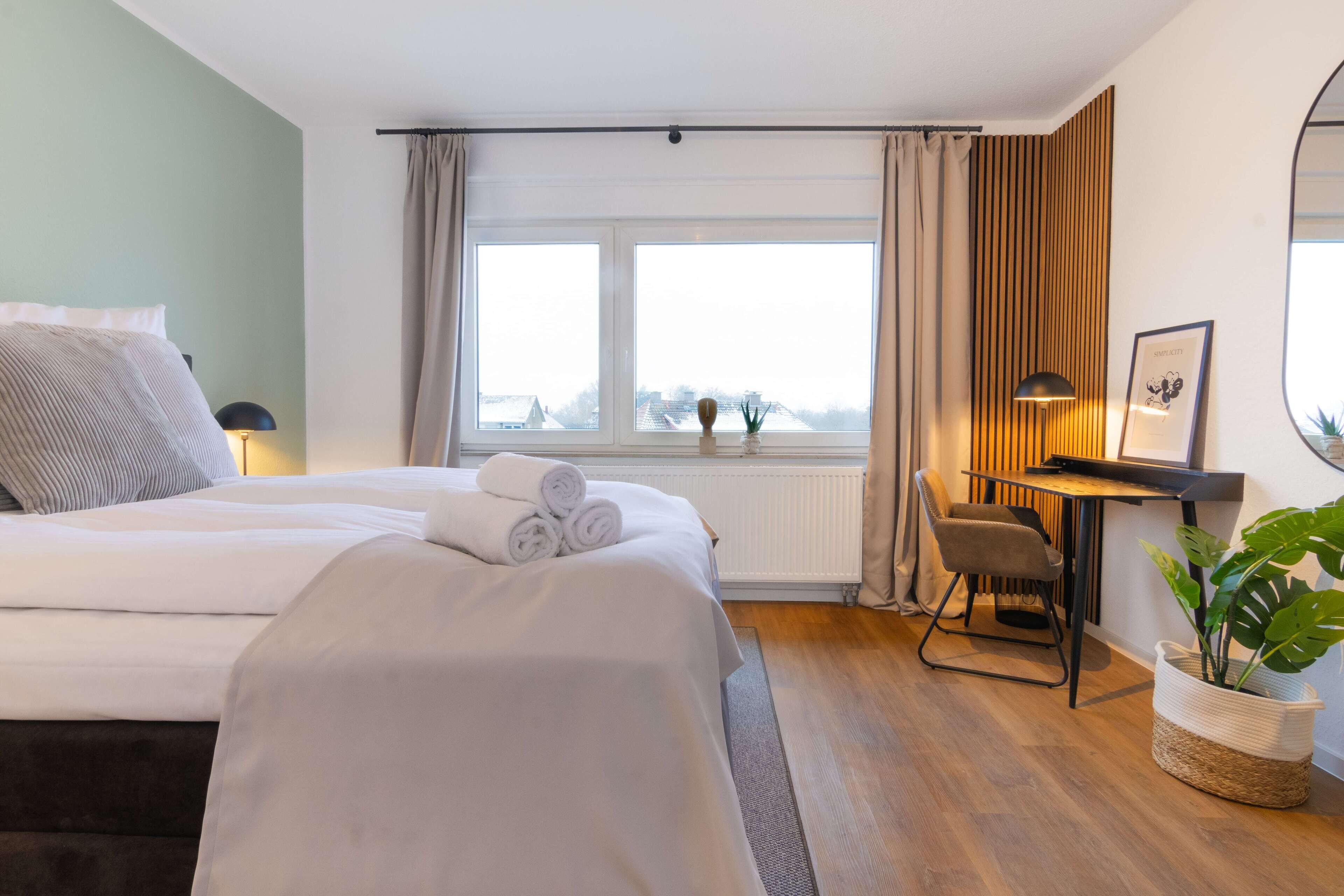 A modern hotel room with a large bed, a desk, a chair, and a view of the city through the window.