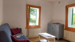 Un espacio habitable compacto con un sofá, una mesita y un armario. Dos ventanas ofrecen luz natural y vistas a la vegetación.