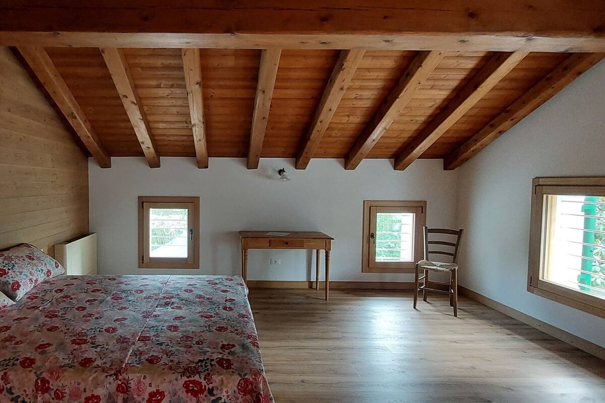 A bedroom with a wooden ceiling, a bed with floral bedding, a desk, and two windows.