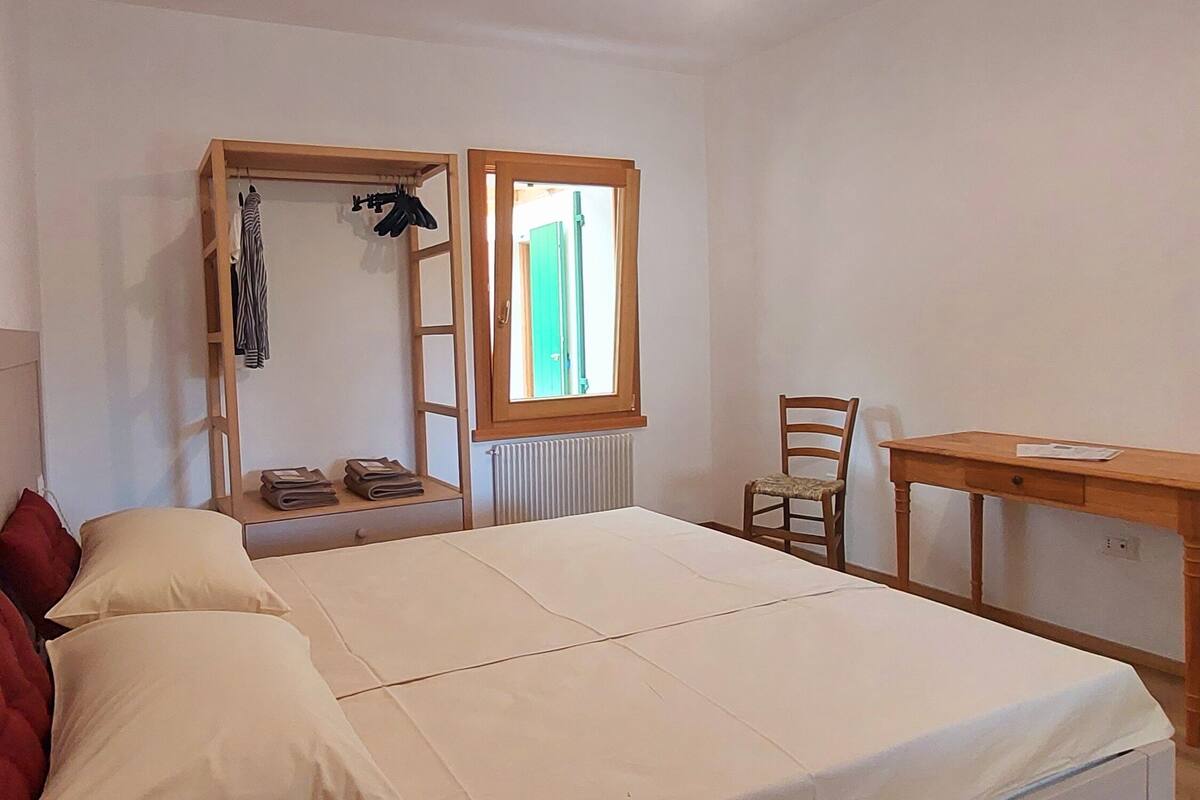 A bedroom with a bed, a wooden desk, a chair, a window with a view, and a hanging light fixture.