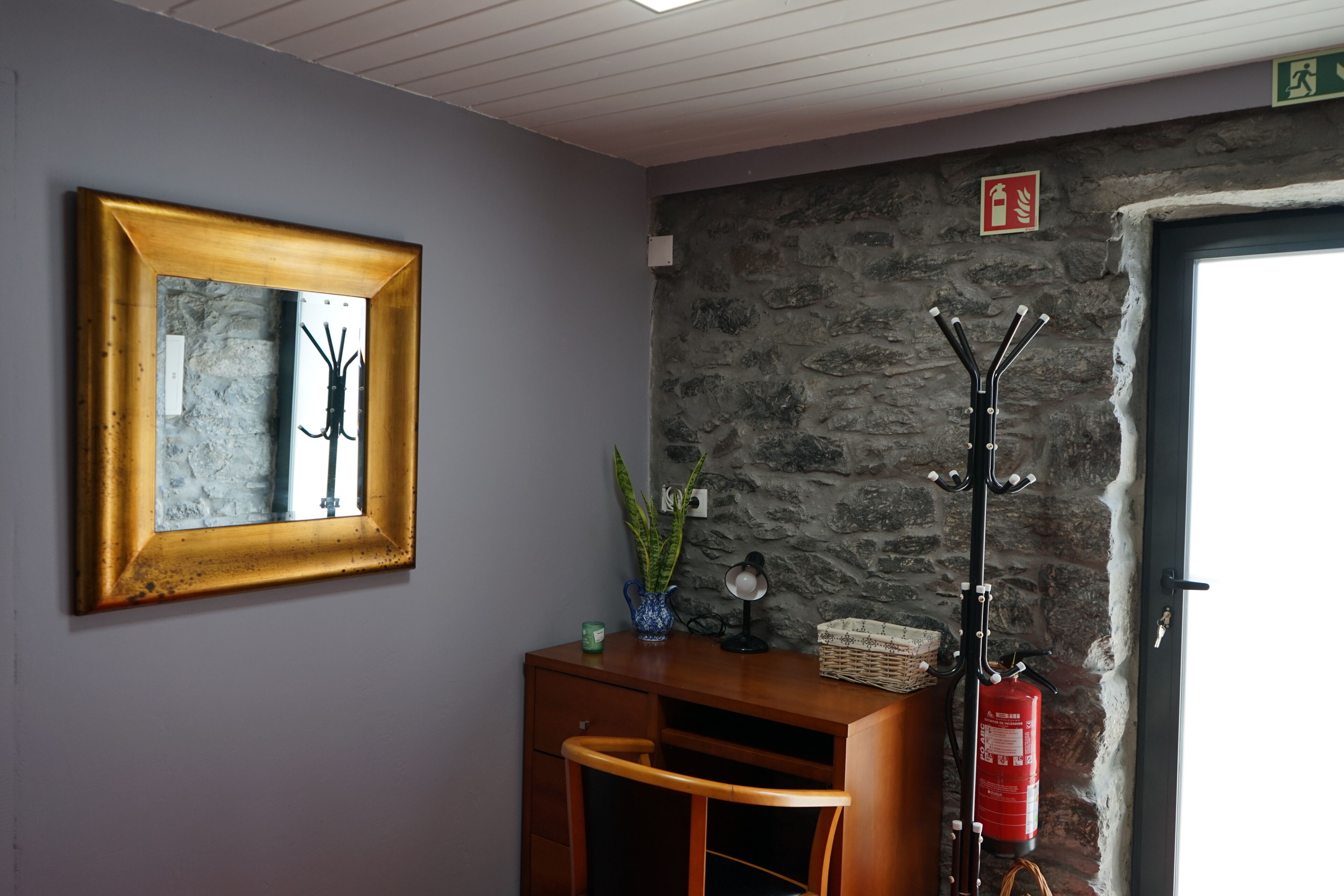 A room with a stone wall, a wooden desk with a lamp, a framed mirror, and a fire extinguisher.