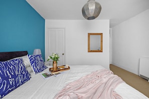 Un lit bien fait, recouvert d’une couverture rose, une table de chevet avec une lampe, un vase rempli de fleurs et un plateau avec de la nourriture.