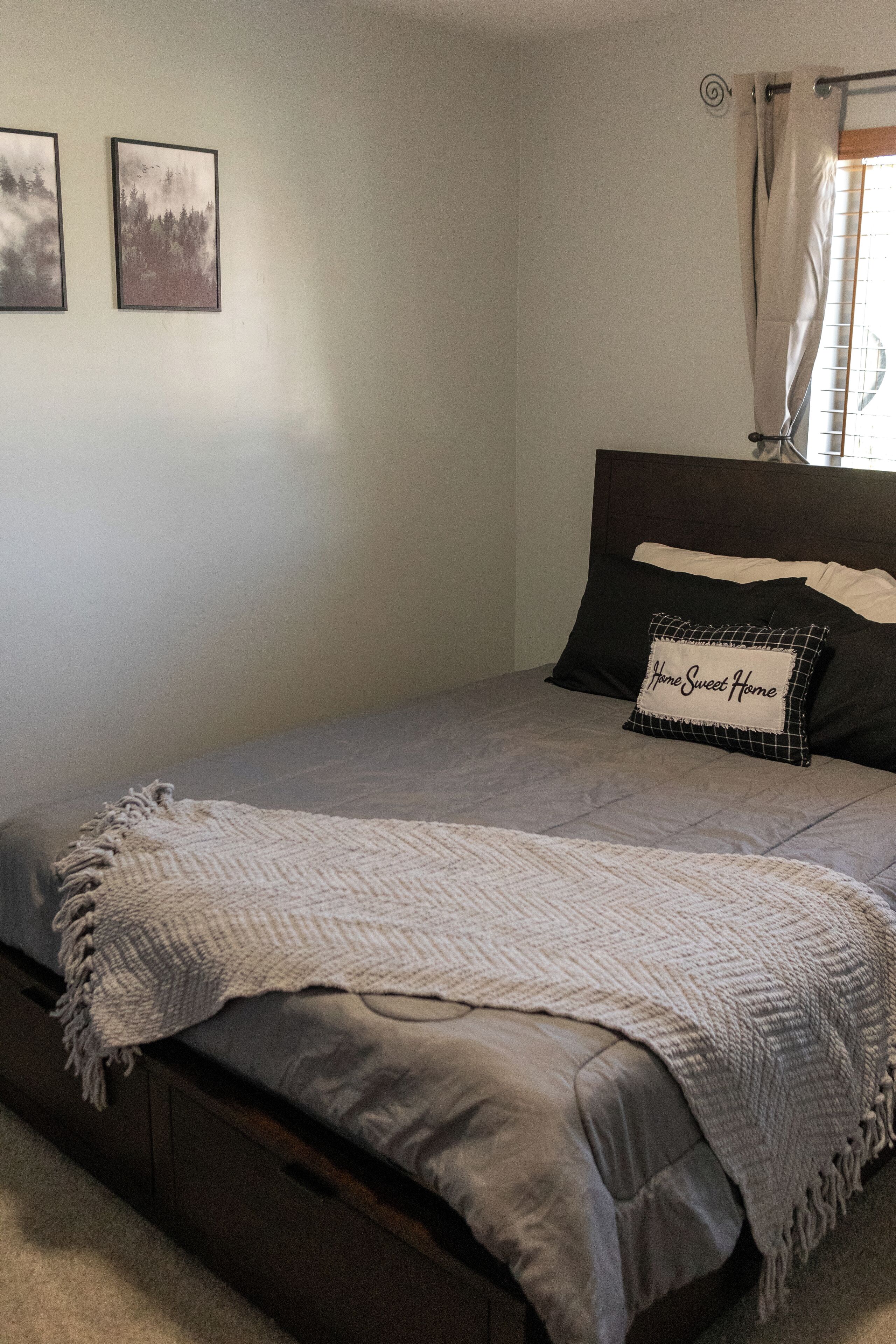 A neatly made bed with a grey blanket, a pillow with "Home Sweet Home" text, and two framed pictures on the wall.
