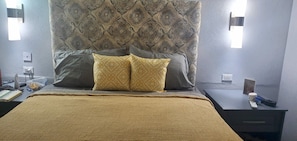 Una cama bien hecha con un cabecero estampado, dos almohadas, una mesita de noche con un control remoto y una lámpara de pared.
