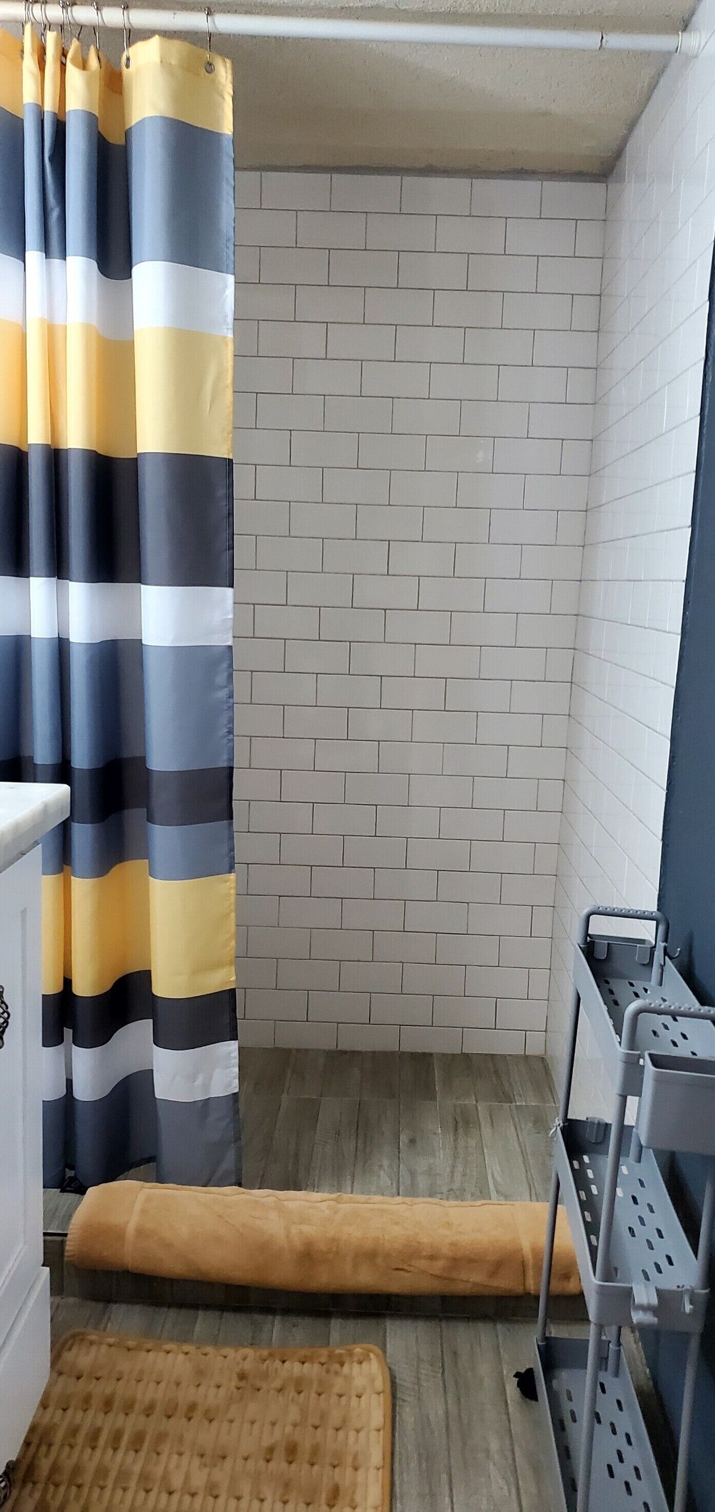 A small bathroom with a shower curtain, a tiled wall, a wooden floor, and a small shelf with a basket.
