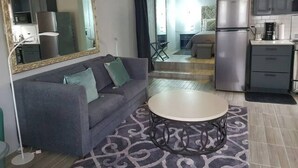 A compact living space with a grey sofa, a round coffee table, a large mirror, and a kitchenette area. - Almond Tree Studio - Modern A/C Apartment w/2 Queen beds in St. James (holetown)