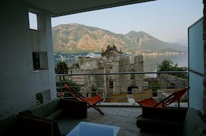 A balcony with a view of mountains and a body of water. There are two chairs and a table on the balcony.