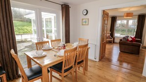A dining area with a wooden table and chairs, a clock on the wall, and a view of the outdoors through large windows. - Gunn's Cottage (Enniskillen)