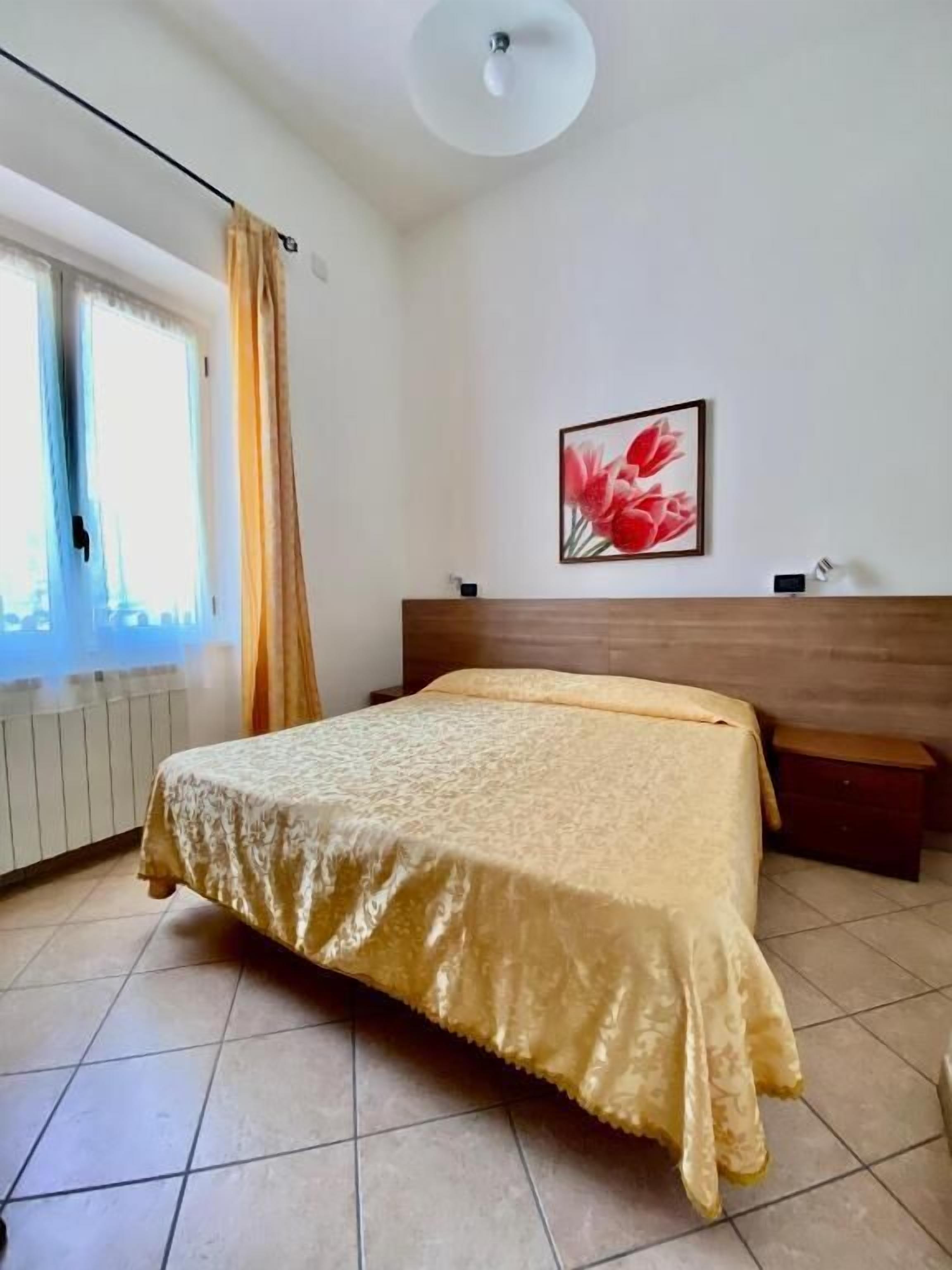 Une chambre à coucher comprenant un lit, une table de chevet, une lampe fixée au mur, une fenêtre avec des rideaux et un tableau encadré accroché au mur.