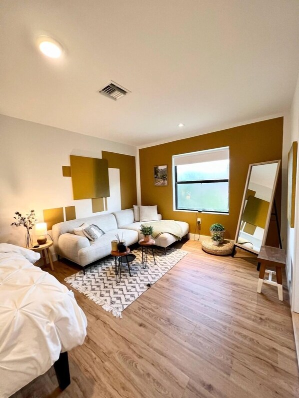 A modern bedroom with a sofa bed, a small round table, a standing mirror, and a large window.
