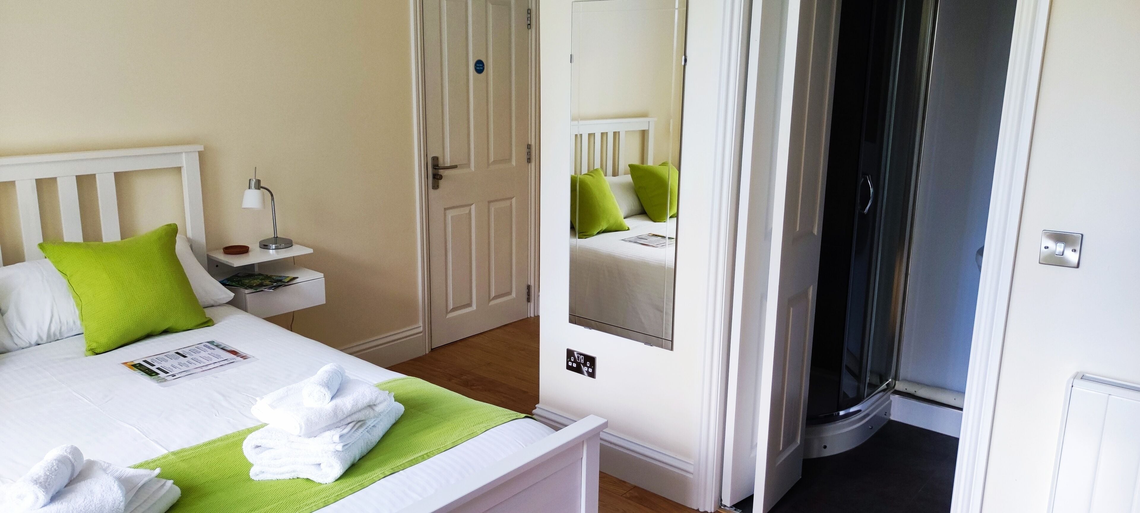 A neatly made bed with white bedding and green pillows, a bedside table with a lamp, and a door leading to a bathroom.
