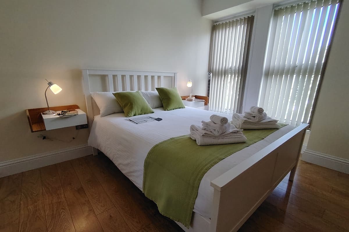 Ein ordentlich bezogenes Bett mit weißem Kopfteil, grünen Kissen und einer grünen Tagesdecke. Zwei Nachttische mit Lampen stehen bereit, und Handtücher sind sauber auf dem Bett gefaltet.