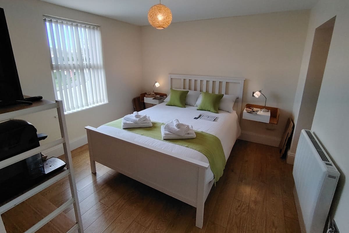 Ein ordentlich eingerichtetes Schlafzimmer mit Bett, Nachttischen, einer Lampe, einem Heizkörper und einem Fenster mit Jalousien.