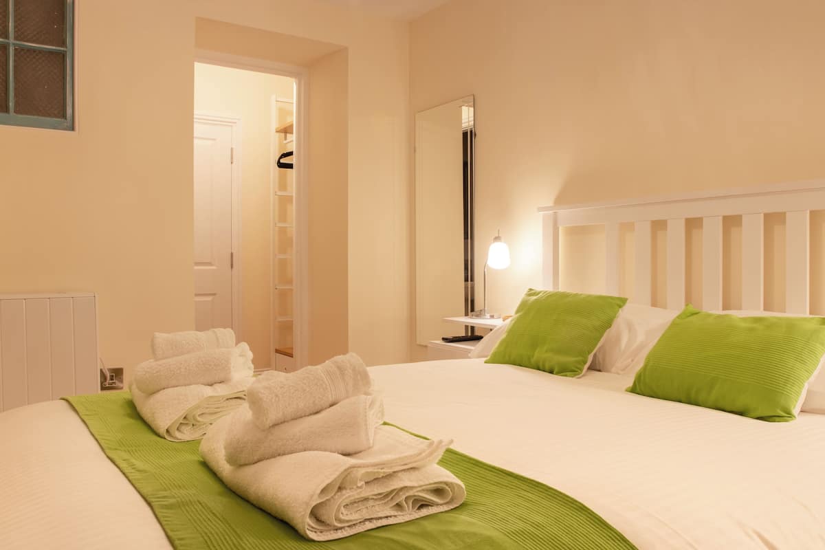 Ein Hotelzimmer mit einem ordentlich bezogenen Bett, einem Nachttisch mit Lampe, einem Spiegel und einer grünen Tagesdecke.