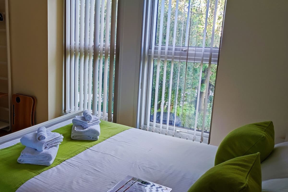 Ein Hotelzimmer mit Bett, Fernseher auf einem Regal, Schreibtisch mit Computer und Fenster mit Jalousien.