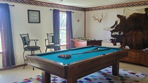 Une table de billard dans une pièce où se trouvent un grand tapis en fourrure, une table de baby-foot et deux chaises.