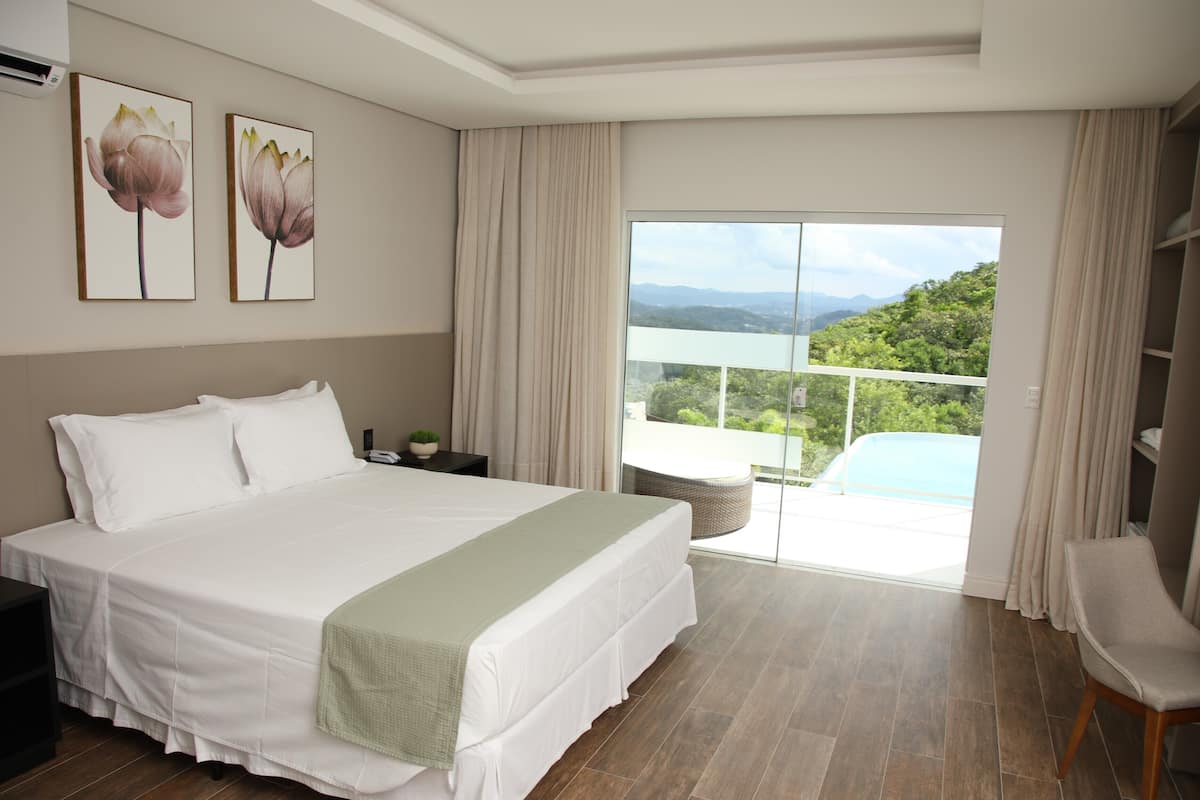 Quarto com uma cama grande, mesa de cabeceira, cadeira e vista para uma varanda com piscina.