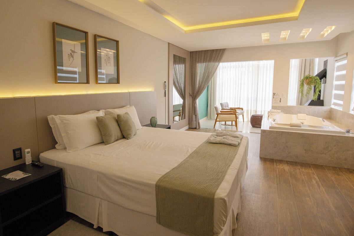 Quarto de hotel moderno com uma cama grande, área de estar com sofá e cadeiras, banheiro separado com banheira e obras de arte nas paredes.