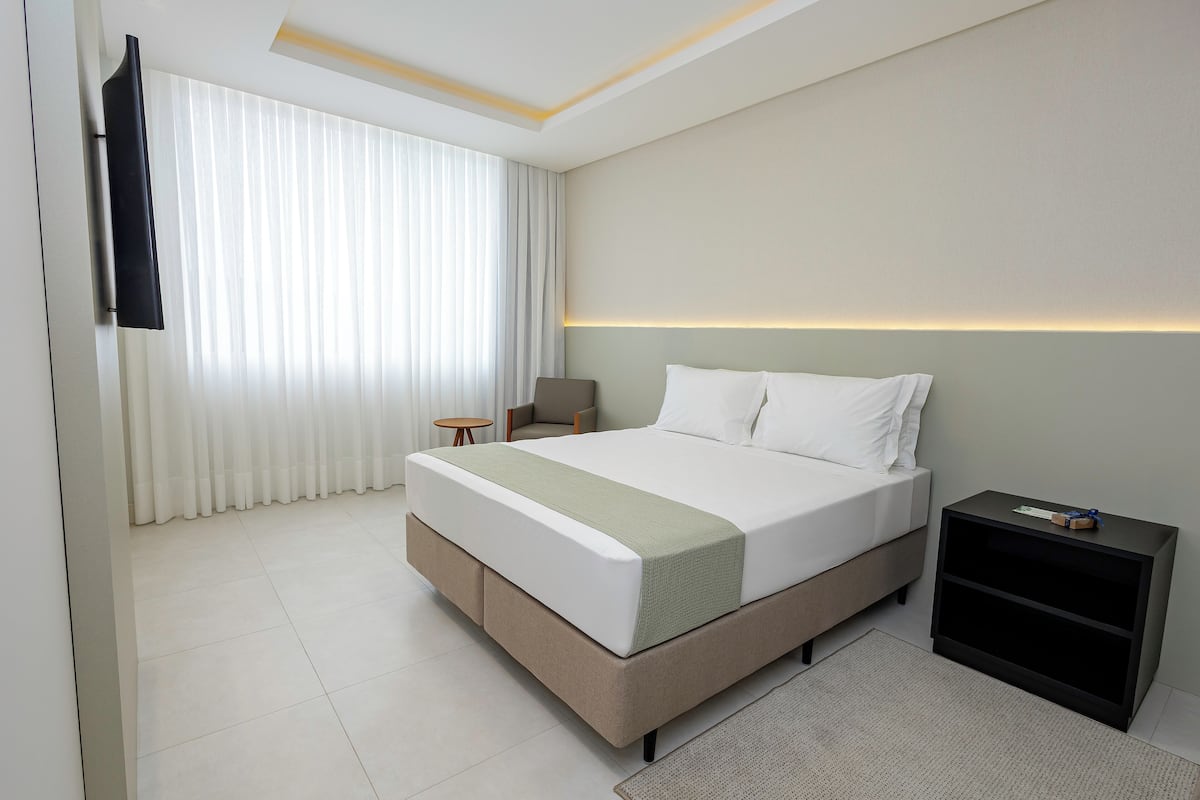 Quarto de hotel moderno com uma cama grande, mesa de cabeceira, televisão montada na parede e janela com cortinas leves.